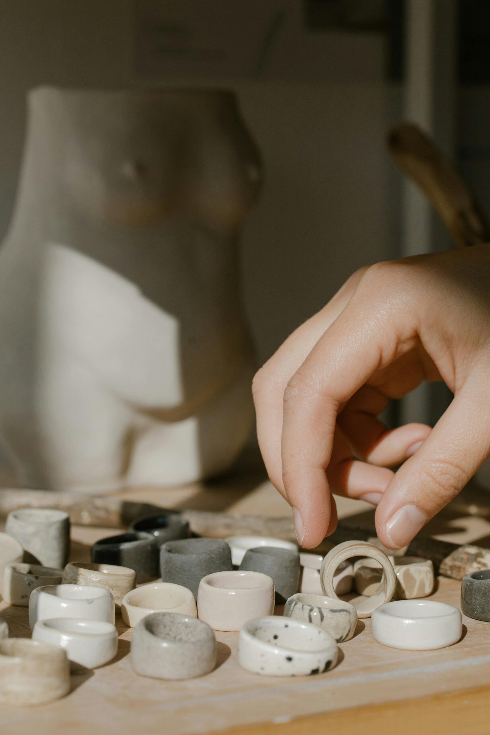 A close-up view of a hand choosing unique handmade ceramic rings on a wooden surface with artistic decor in the background.